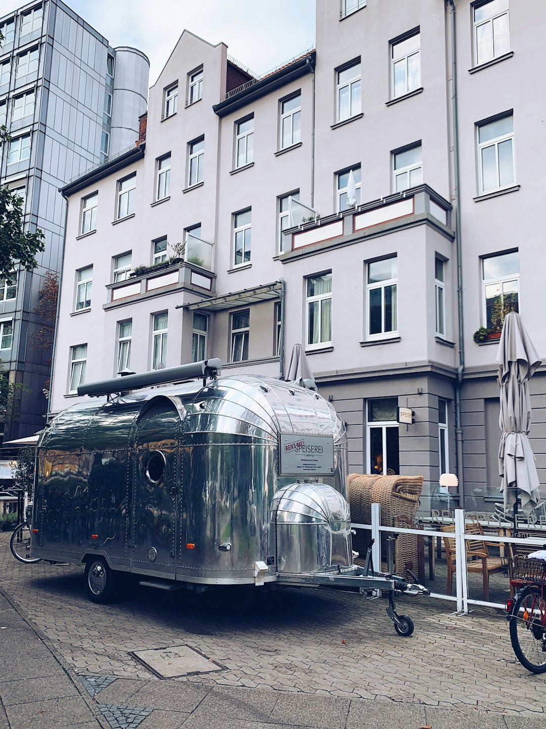 Silver food trailer parked on a cobblestone street near an outdoor cafe.